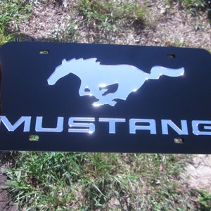 Puede incluir: Placa de matr&iacute;cula de metal negro con un logotipo de Mustang plateado y la palabra "MUSTANG" en letras plateadas.