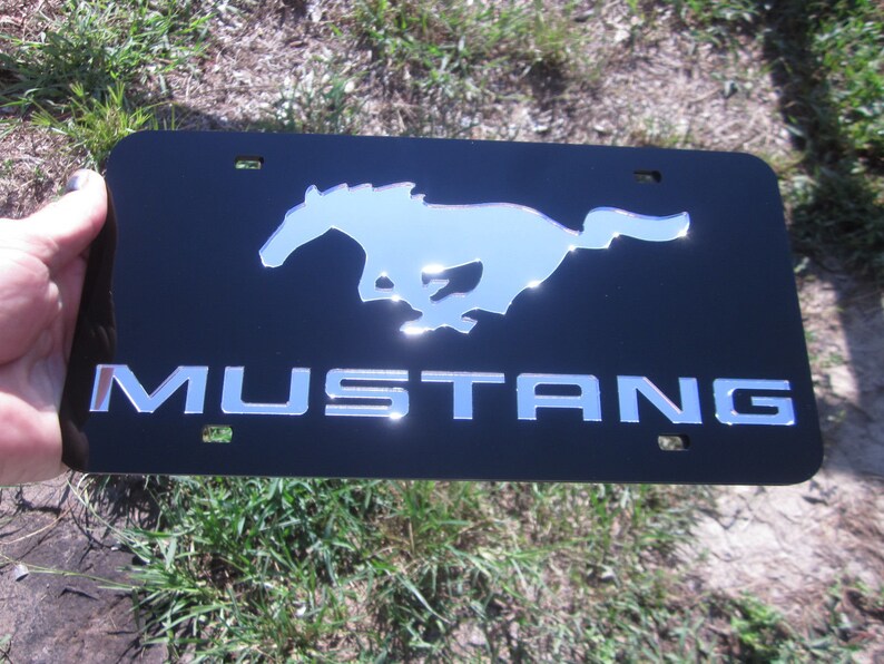 Puede incluir: Placa de matr&iacute;cula de metal negro con un logotipo de Mustang plateado y la palabra "MUSTANG" en letras plateadas.