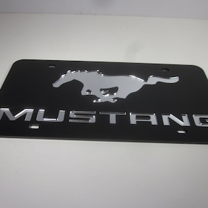 Puede incluir: Marco de matr&iacute;cula negro con un logotipo de Mustang en cromo plateado y la palabra "MUSTANG" en letras de cromo plateado.