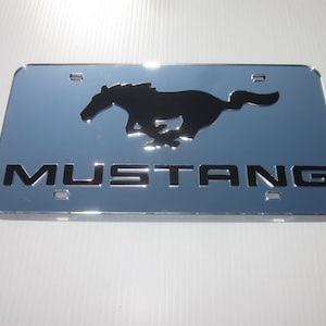 Puede incluir: Un marco de matr&iacute;cula cromado con una silueta negra de un caballo corriendo y la palabra "MUSTANG" en letras negras.