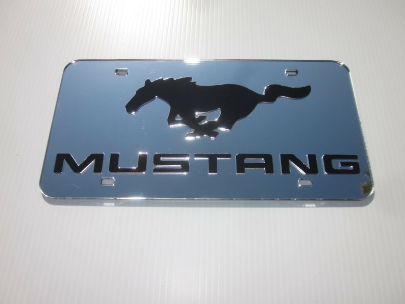 Puede incluir: Un marco de matr&iacute;cula cromado con una silueta negra de un caballo corriendo y la palabra "MUSTANG" en letras negras.