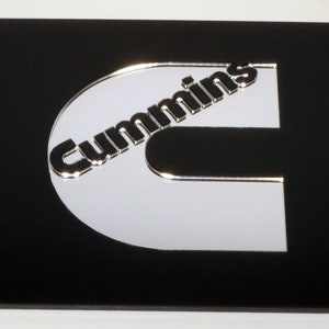 May include: Black license plate with a chrome Cummins logo. The logo is a stylized letter C with the word "Cummins" in a curved font. The plate has four mounting holes.