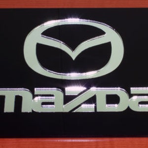 Puede incluir: Placa de matrícula negra con un logotipo de Mazda plateado y la palabra "Mazda" en letras plateadas.