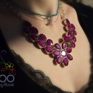 May include: A silver necklace with a large pink flower pendant made of oval-shaped gemstones. The necklace is layered over a silver chain and a turquoise beaded necklace.