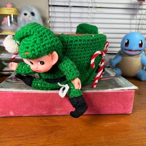May include: A handmade Christmas elf doll with a green crocheted outfit, including a hat and a bag. The elf has a red and white candy cane and is sitting on a red box. A blue Squirtle figure is in the background.
