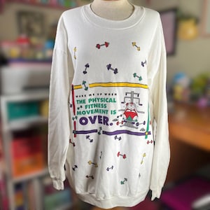 May include: White long-sleeve sweatshirt with a colorful graphic design. The design includes scattered dumbbells and the text "WAKE ME UP WHEN THE PHYSICAL FITNESS MOVEMENT IS OVER." A person is depicted using a weight machine. The sweatshirt is on a mannequin.