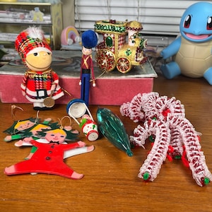 May include: A collection of Christmas ornaments and decorations, including a toy train, a soldier, a doll, and a candy cane-shaped decoration. The ornaments are in various colors, including red, green, gold, and white. The items are arranged on a wooden surface.