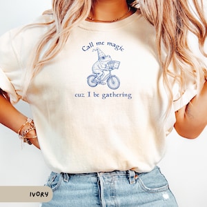 May include: Ivory-colored sweatshirt with a blue illustration of a frog riding a bicycle, wearing a wizard hat, and carrying a basket. The text "Call me magic cuz I be gathering" is above and below the frog.