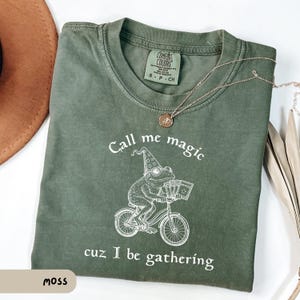 May include: Moss green t-shirt with the text "Call me magic cuz I be gathering" and a graphic of a frog riding a bicycle and wearing a wizard hat. A gold necklace with a pendant is on the shirt.