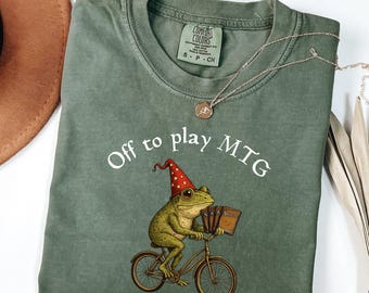 May include: Moss green t-shirt with the text "Off to play MTG" and a frog riding a bicycle, wearing a wizard hat and holding cards. A silver necklace and a brown hat are also visible.
