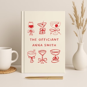 May include: A cream-colored hardcover notebook featuring red line-art illustrations of six different cocktail glasses, each adorned with a small bow. The text "THE OFFICIANT" and "ANNA SMITH" is printed in red in the center of the front cover.