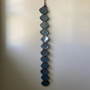 May include: A vertical wall hanging composed of ten blue-grey, fan-shaped ceramic pieces. The pieces are strung together with a brown leather cord, creating a decorative element. The wall hanging is mounted on a neutral-colored wall.