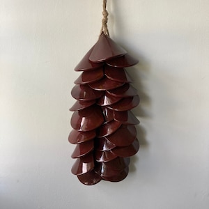 May include: A decorative wall hanging resembling a pine cone, crafted from multiple, overlapping, dark red, cone-shaped pieces. The pieces are arranged in a cascading pattern and suspended from a natural fiber rope.