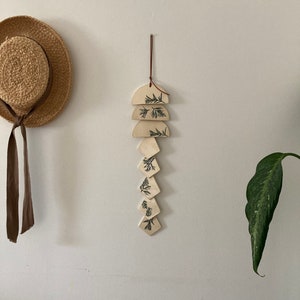 May include: A white ceramic wall hanging with a brown leather cord. The hanging features a series of geometric shapes, including half circles and diamonds, with a leafy pattern in black.