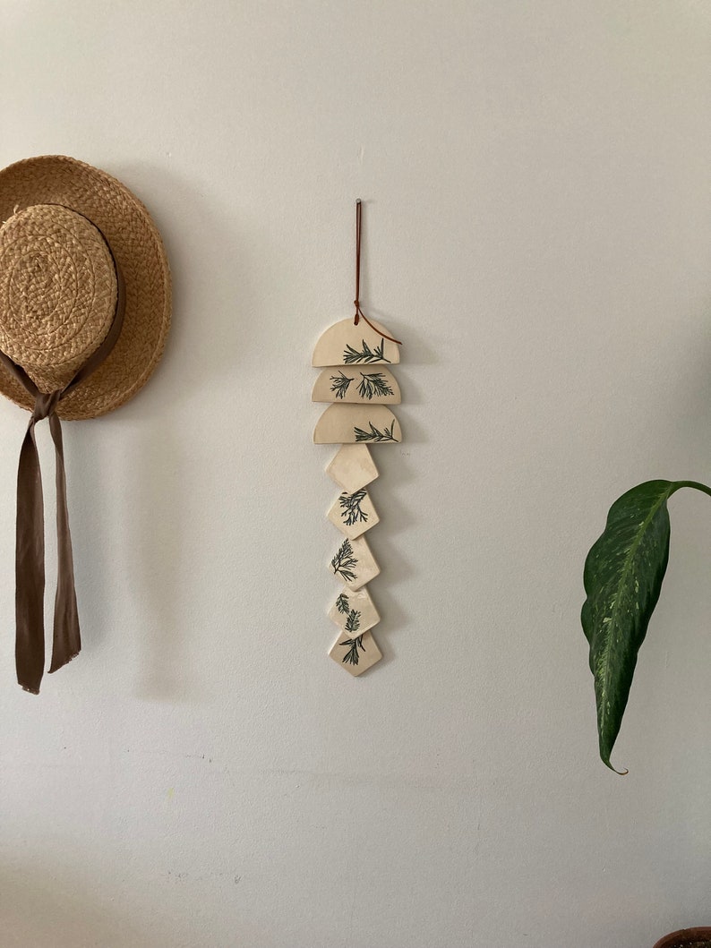 May include: A white ceramic wall hanging with a brown leather cord. The hanging features a series of geometric shapes, including half circles and diamonds, with a leafy pattern in black.