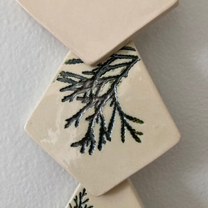 May include: Three white ceramic tiles with a diamond shape. Each tile has a black and green leafy design.