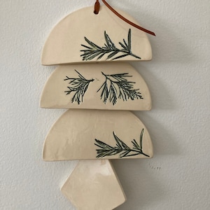 May include: A white ceramic wall hanging with a brown leather cord. The hanging features four pieces in the shape of half circles and a diamond. Each piece has a black line drawing of a sprig of greenery.