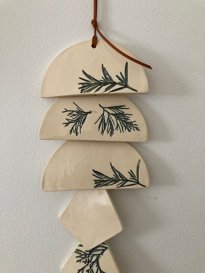 May include: A white ceramic wall hanging with a brown leather cord. The hanging features four pieces in the shape of half circles and a diamond. Each piece has a black line drawing of a sprig of greenery.