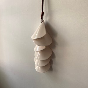 May include: A white ceramic mobile with a brown leather cord. The mobile is made up of six overlapping, cone-shaped pieces.