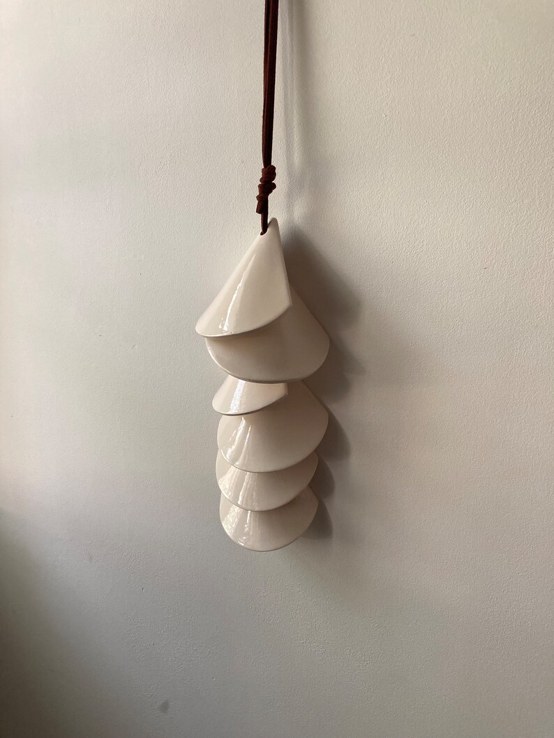 May include: A white ceramic mobile with a brown leather cord. The mobile is made up of six overlapping, cone-shaped pieces.
