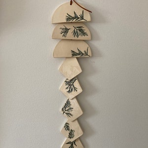 May include: A white ceramic wind chime with a brown leather cord. The chime features a series of semi-circular and diamond-shaped pieces with a green leaf design.