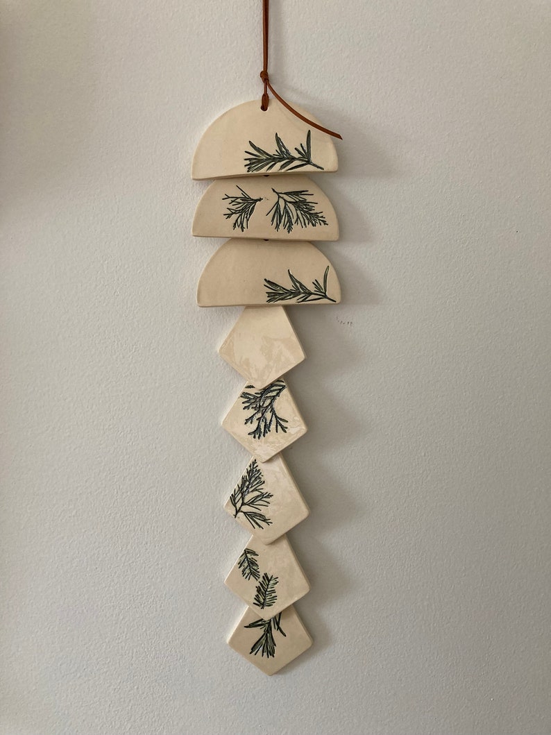 May include: A white ceramic wind chime with a brown leather cord. The chime features a series of semi-circular and diamond-shaped pieces with a green leaf design.
