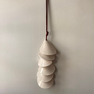 May include: A white ceramic wind chime with a brown leather cord hanging from the top. The chime is made up of seven overlapping, cone-shaped pieces.