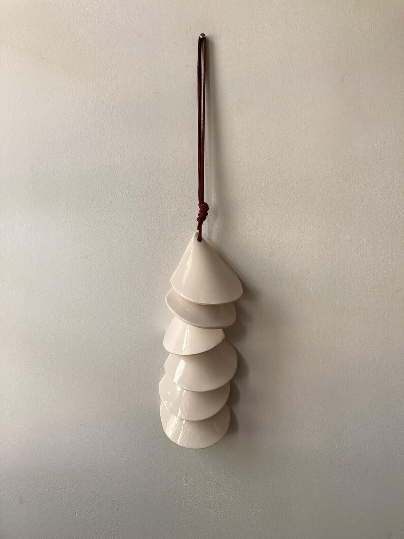 May include: A white ceramic wind chime with a brown leather cord hanging from the top. The chime is made up of seven overlapping, cone-shaped pieces.