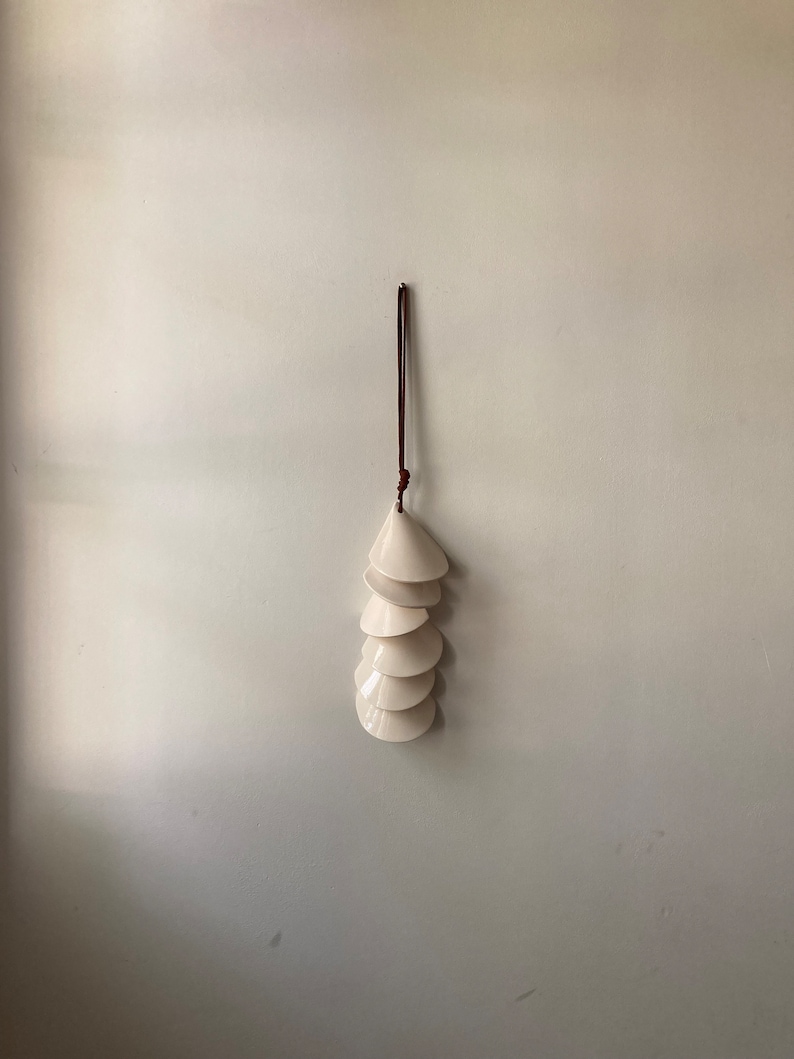 May include: A white ceramic wind chime with a brown leather cord hanging from the ceiling. The chime is made up of seven overlapping, cone-shaped pieces.