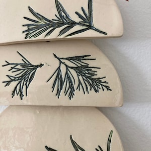 May include: Three ceramic ornaments in the shape of half circles, each with a different black line drawing of a sprig of rosemary.