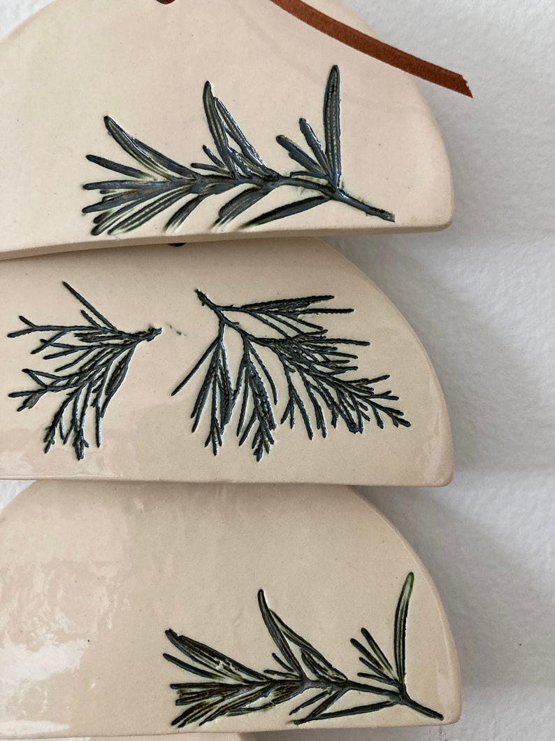May include: Three ceramic ornaments in the shape of half circles, each with a different black line drawing of a sprig of rosemary.