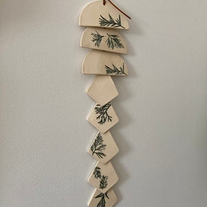 May include: A white ceramic wind chime with a brown leather cord and green leaf designs. The chime is made up of six pieces, three semi-circles and three diamonds.