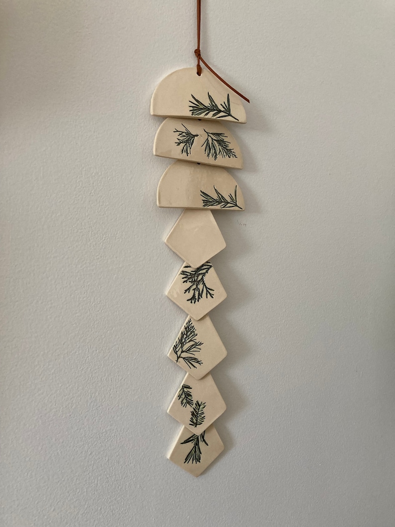 May include: A white ceramic wind chime with a brown leather cord and green leaf designs. The chime is made up of six pieces, three semi-circles and three diamonds.