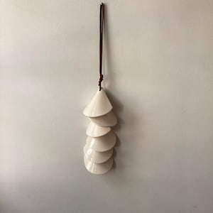 May include: A white ceramic wind chime with a brown leather cord hanging from the ceiling. The chime is made up of seven cone-shaped pieces stacked on top of each other.