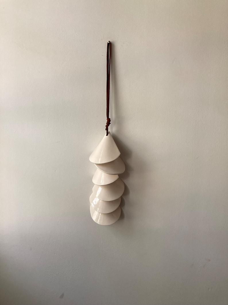 May include: A white ceramic wind chime with a brown leather cord hanging from the ceiling. The chime is made up of seven cone-shaped pieces stacked on top of each other.