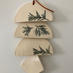 May include: A white ceramic wall hanging with a brown leather cord. The hanging features four pieces in the shape of half circles and a diamond. Each piece has a black line drawing of a sprig of greenery.
