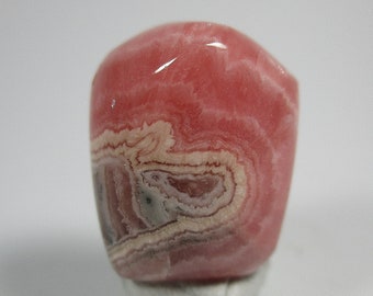 May include: A polished, rounded gemstone with a vibrant pink hue, featuring swirling patterns of white, gray, and darker pink. The stone is set on a small, light-colored base, showcasing its natural beauty.