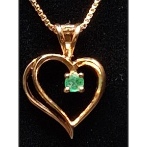 May include: A gold-colored pendant necklace featuring a heart-shaped design. The pendant has a small, square-cut emerald gemstone at its center. The necklace chain is also gold-colored, creating a cohesive and elegant look.
