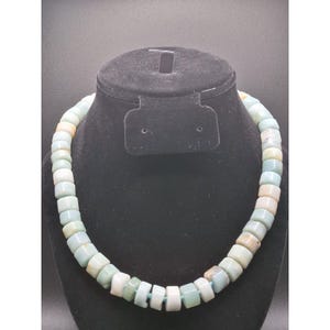 May include: A necklace composed of rectangular, multi-colored beads in shades of blue, green, and peach. The beads are strung together, creating a chunky, eye-catching accessory. The necklace is displayed on a black mannequin bust against a dark background.
