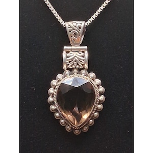 May include: A silver pendant necklace featuring a pear-shaped, faceted brown gemstone set in a beaded frame. The pendant is attached to a silver box chain by a decorative, engraved bail with intricate scrollwork patterns and a hinged design.