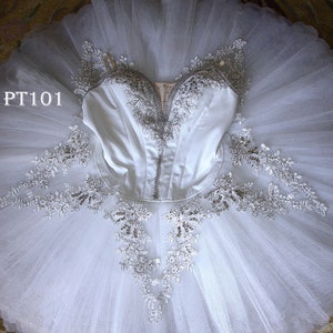 Może przedstawiać: Biały tiulowy tutu baletowy z białym gorsetem i srebrnymi ozdobami. Gorset ma koronkową lamówkę, a tutu ma warstwowy design. Tutu jest oznaczone "PT101".
