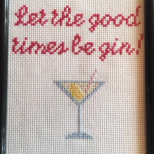 Könnte beinhalten: Kreuzstich-Stickerei in einem schwarzen Rahmen mit weißem Stoff. Die Stickerei zeigt den Text "Let the good times be gin!" in rotem Garn und ein blaues Martini-Glas mit einer gelben Zitronenscheibe und einem rosa Strohhalm.
