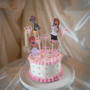 Pode incluir: Um bolo redondo branco decorado com glacê rosa e granulado em forma de coração. O bolo é coberto com enfeites rosa em forma de coração e recortes de personagens de desenhos animados. O bolo está em um prato branco.