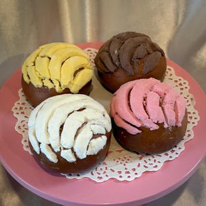 Könnte beinhalten: Vier bunte Concha-Gebäckstücke auf einem rosa Teller mit einer weißen Spitze. Die Conchas haben eine braune Basis mit gelben, weißen, rosa und braunen gemusterten Belägen. Ein mexikanisches Brot.