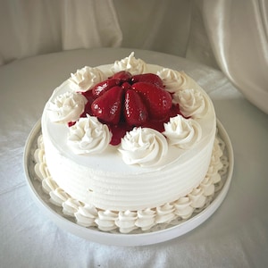 Puede incluir: Un pastel redondo cubierto de glaseado blanco liso con un borde decorativo hecho con manga pastelera en la base y en la parte superior. El centro está decorado con fresas rojas brillantes cubiertas de un glaseado rojo intenso.
