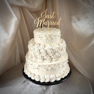 May include: A three-tiered white cake decorated with white frosting rosettes and scattered white pearls. The top tier features a gold-coloured cake topper with the text Just Married in a cursive script font, positioned on a black circular stand.