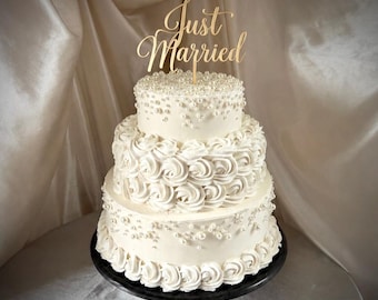 May include: A three-tiered white cake decorated with white frosting rosettes and scattered white pearls. The top tier features a gold-colored cake topper with the text Just Married in a cursive script font, positioned on a black circular stand.