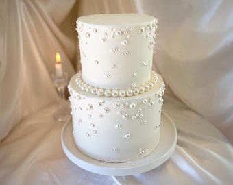 May include: A two-tier white cake decorated with scattered white pearl-like spheres of varying sizes. A single row of larger white pearls encircles the base of the top tier. The cake sits on a plain white circular stand, presenting a smooth finish.
