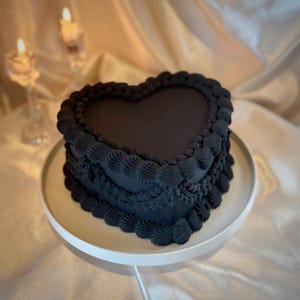 Puede incluir: Un pastel en forma de corazón con glaseado oscuro, decorado con remolinos de crema de mantequilla. El pastel está sobre un soporte blanco. Dos velas encendidas están en el fondo.