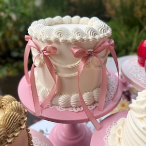 Peut inclure: Un gâteau rond blanc au glaçage lisse, décoré de bordures en crème fouettée sur les bords supérieur et inférieur. Les côtés présentent de délicates guirlandes en crème et sont ornés de plusieurs fins rubans de satin rose poudré.